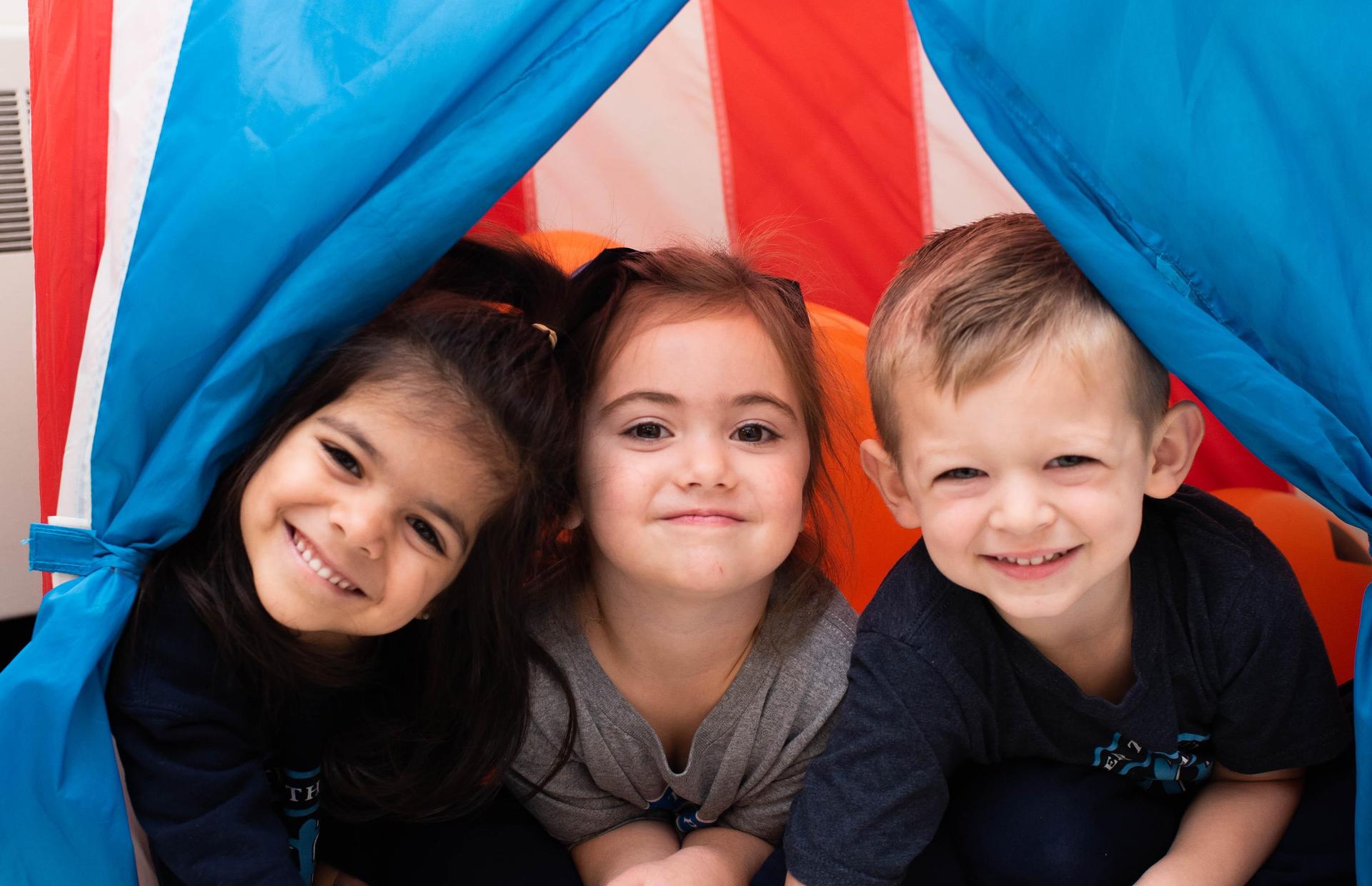 Three children smile while peeking out from a colorful play tent.