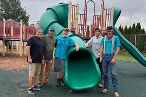 A green sliding board with five men standing alongside on a playground