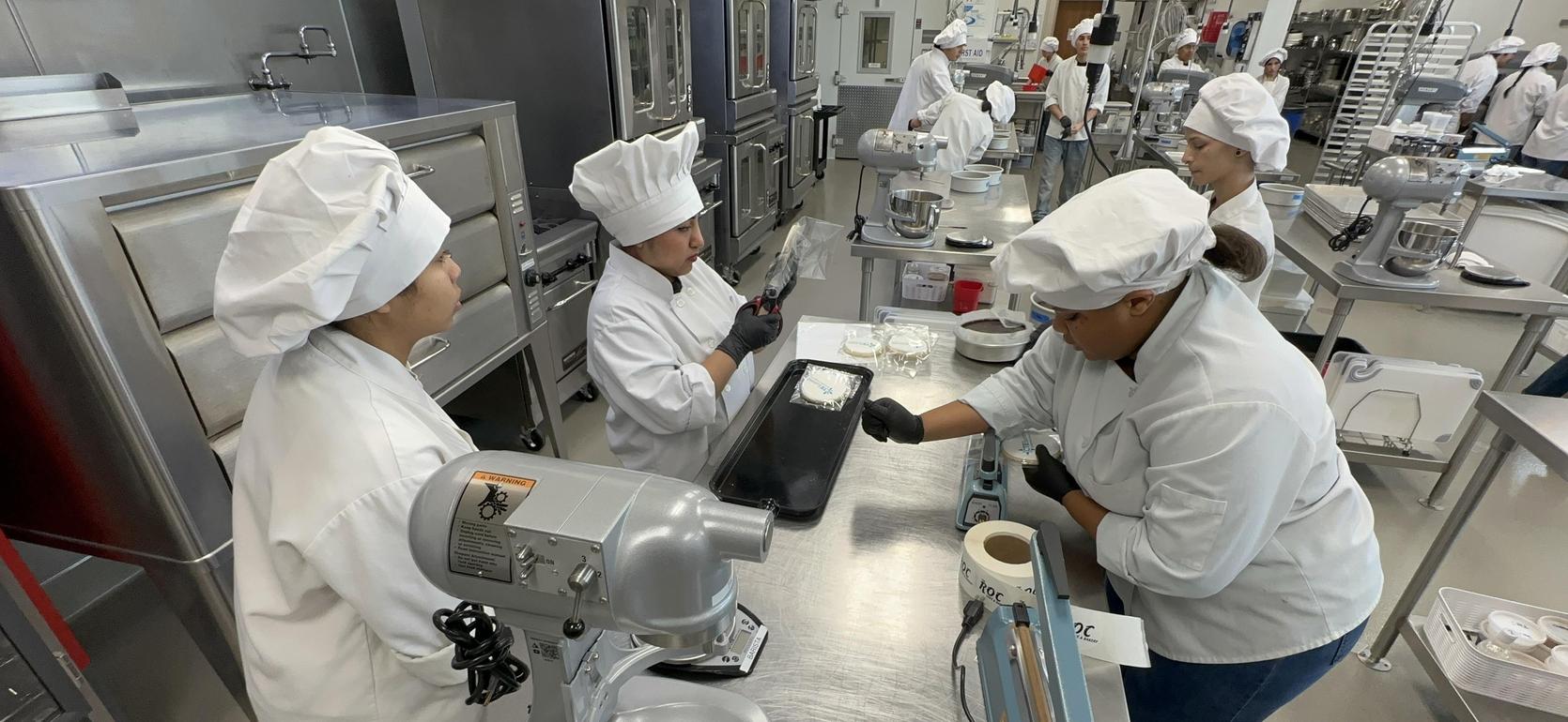 Four chefs in a kitchen working with equipment and ingredients.