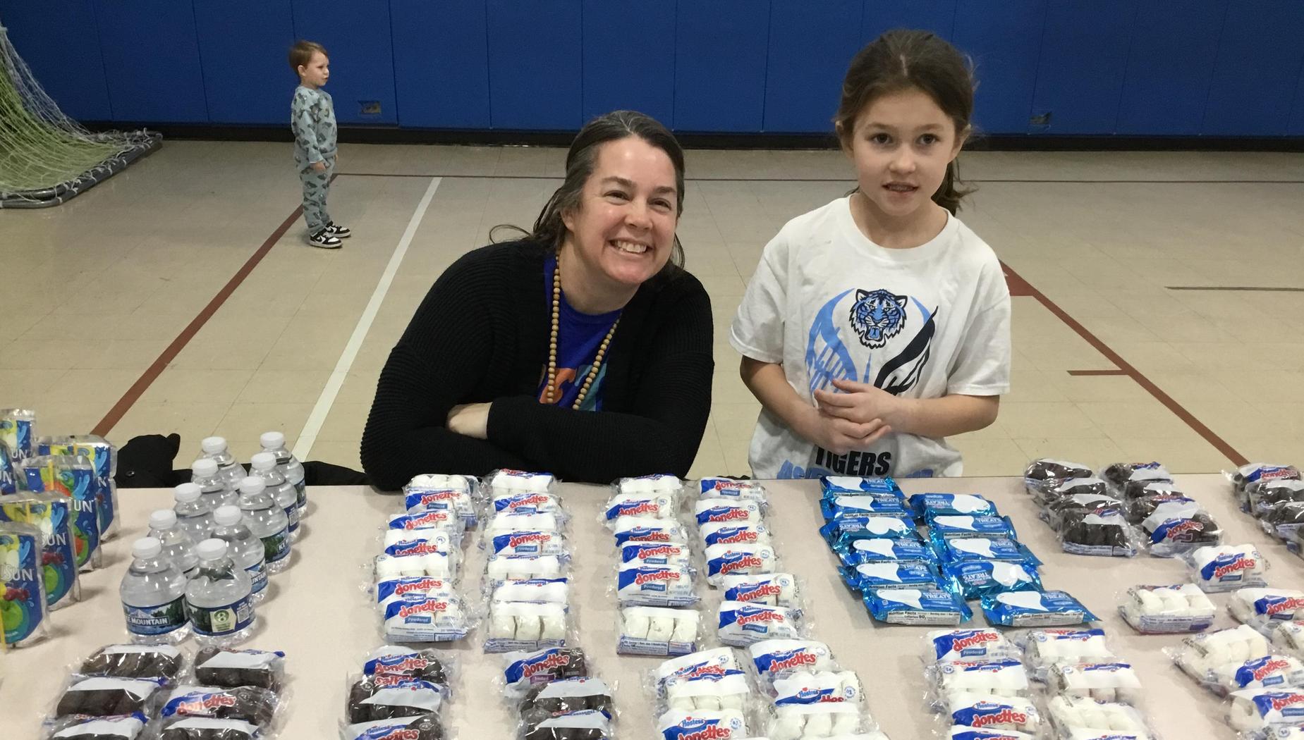 A woman and a girl smile at a table filled with snacks at a school event.