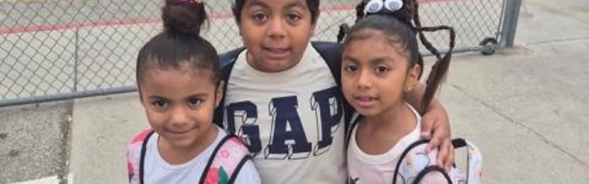 Three children smiling together outside near a fence.