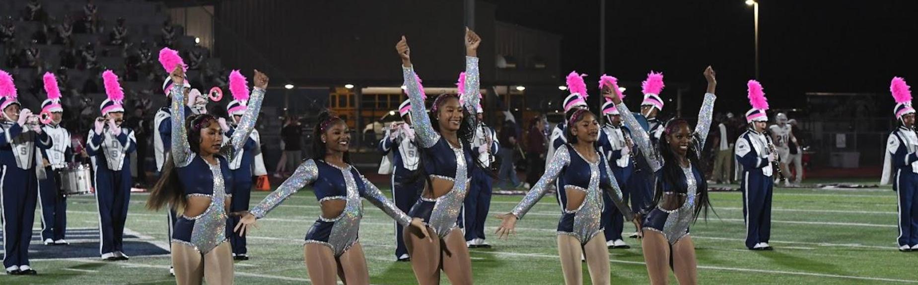 Five performers in sparkling outfits dance joyfully, with a marching band visible in background.
