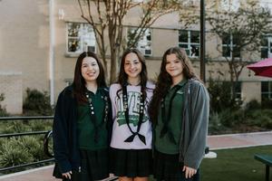 Three girls in matching outfits smiling together outside on a school campus.