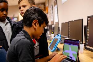 A child interacts with a laptop game while two peers look on.