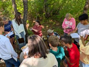 Students gathering around an instructor in a green, wooded area.