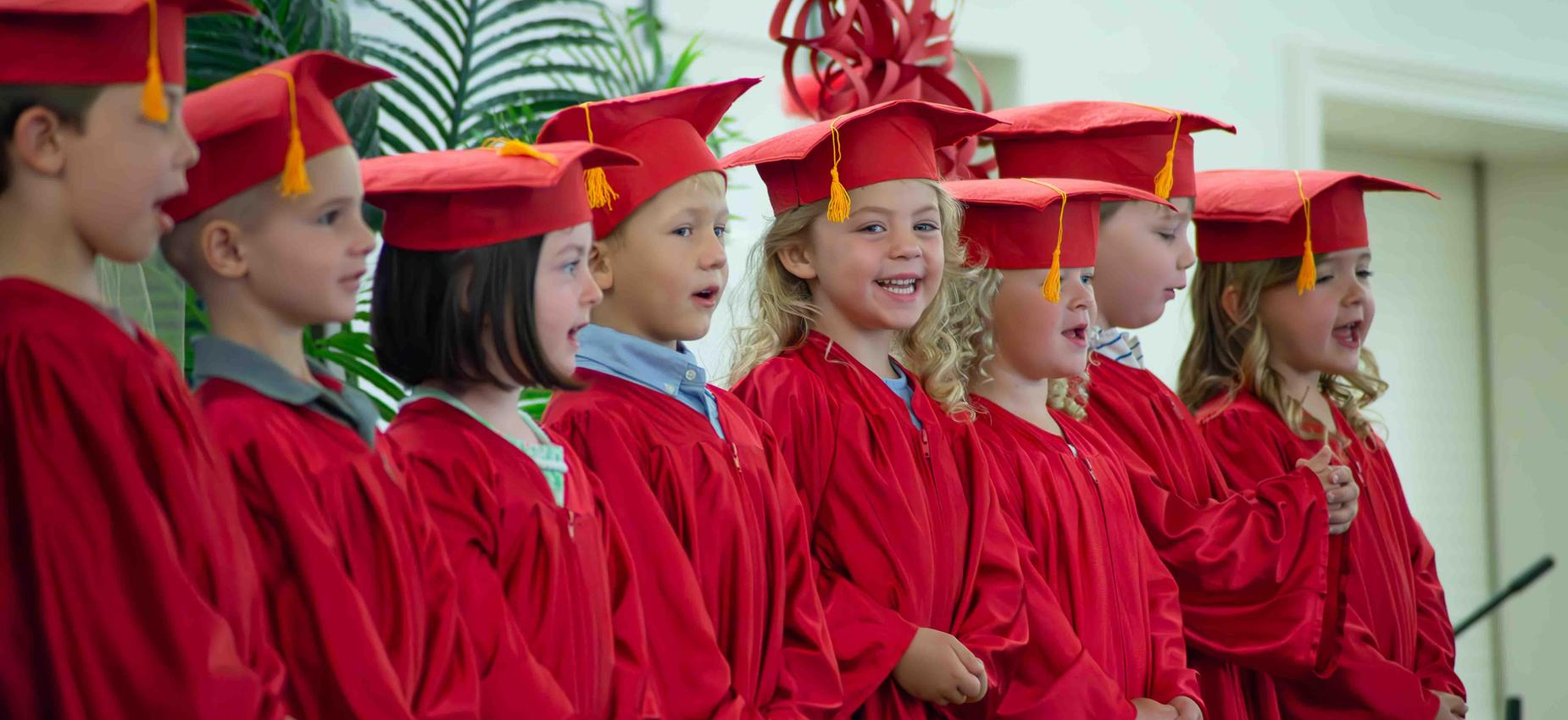 A line of children in red graduation gowns and caps smiling during a ceremony.