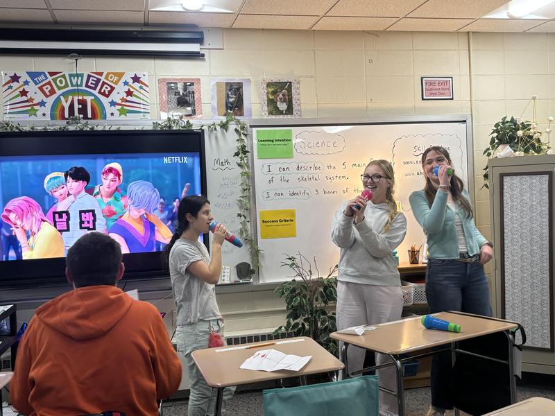 Students singing in class with a large screen displaying a colorful performance.