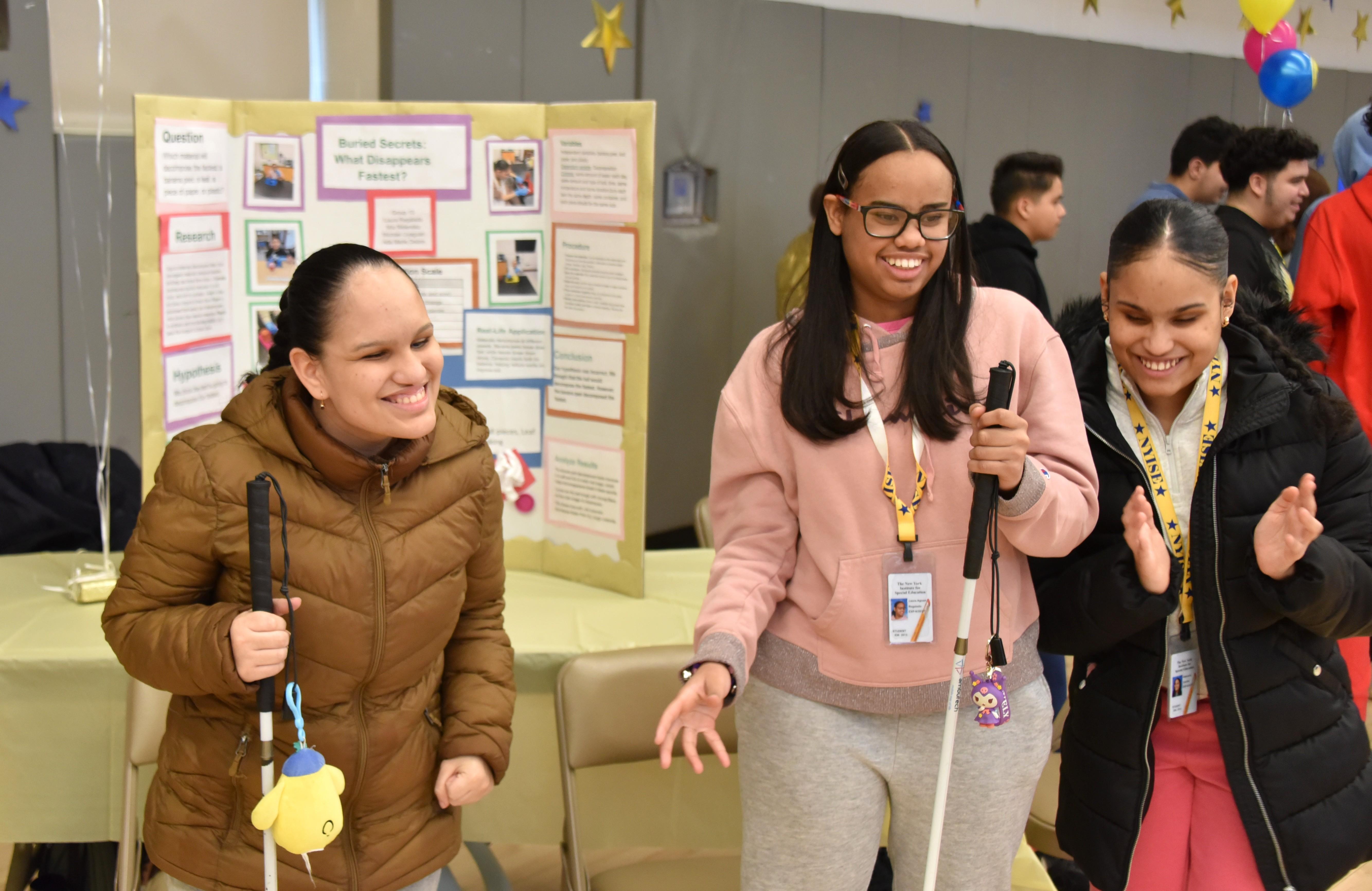 3 girls with white canes are standing in front of a science project in a gymnasium