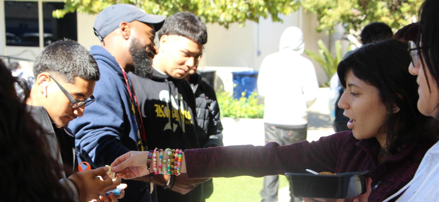 A group of young people interacting outdoors, one handing food to another.