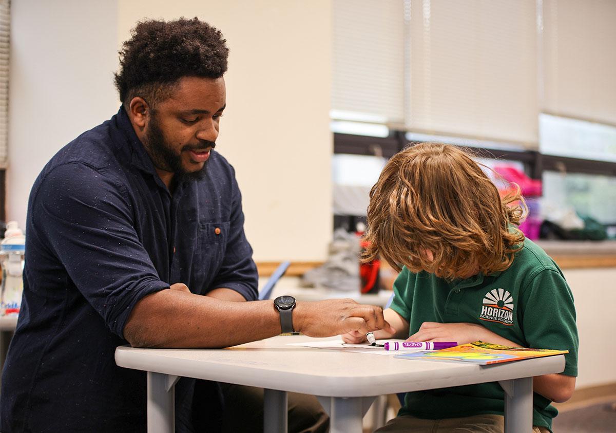 HSA Teacher smiles while kneeling beside a young student in a classroom setting.