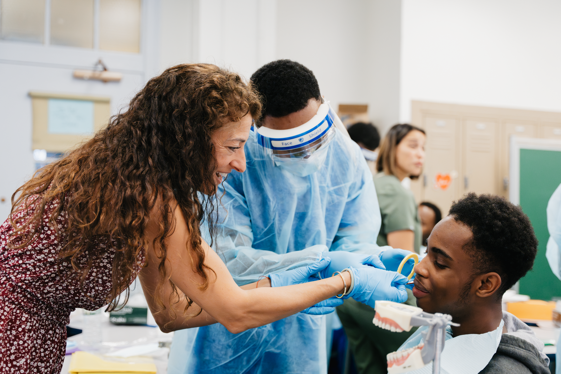Instructor guides a student in protective gear while they practice a dental procedure on a model.