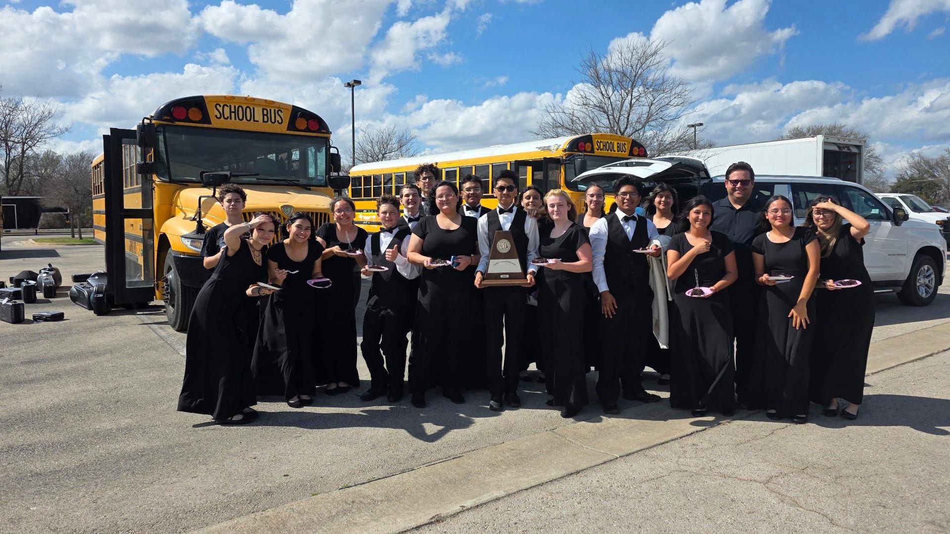 A group of performers in black outfits celebrate with a trophy near school buses.