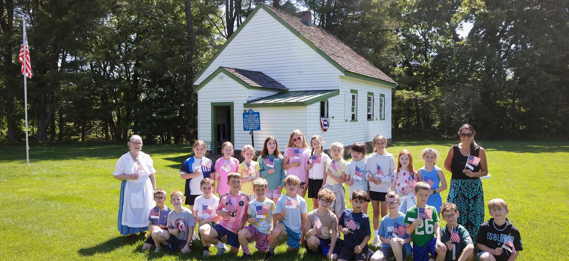 Group of children and adults holding American flags in front of a white schoolhouse.