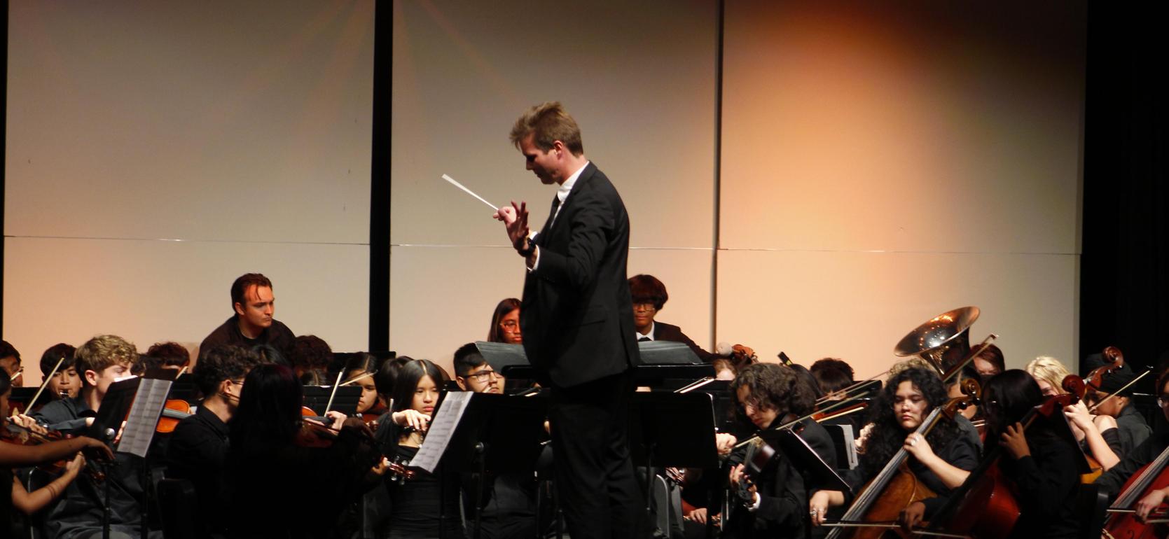 Conductor leading a large orchestra on stage with musicians playing various instruments.