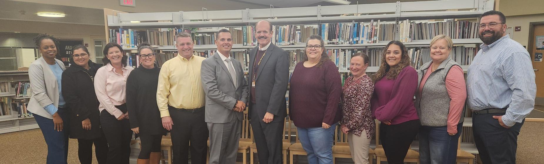 A group of thirteen people standing together in a library setting, smiling and posing for a photo.