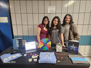 Three individuals pose behind a table displaying items, including a game wheel and a book.