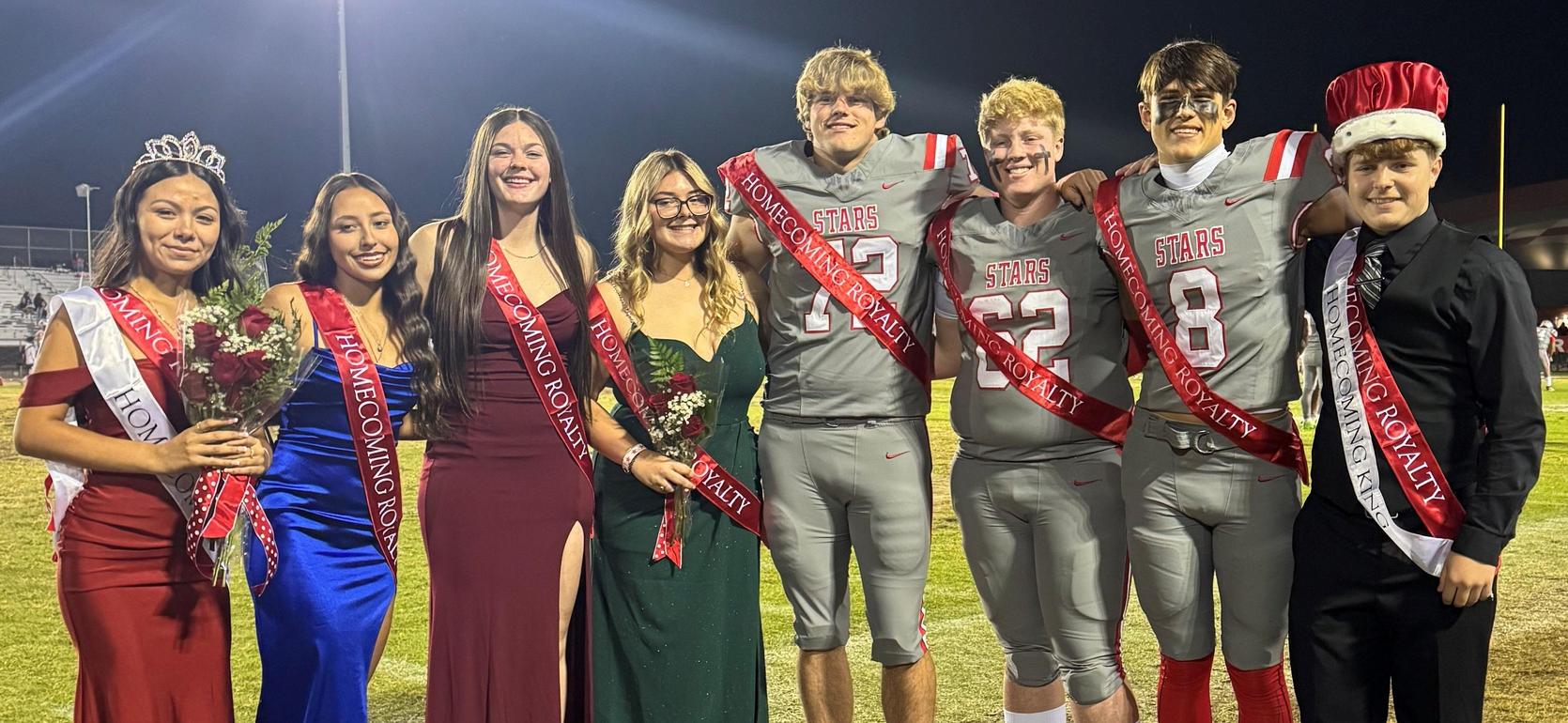 A group of homecoming royalty candidates poses on a football field, wearing sashes and formal attire.