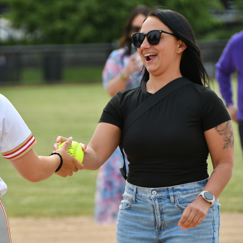A woman in a black shirt smiling while shaking hands with another person, holding a yellow softball.