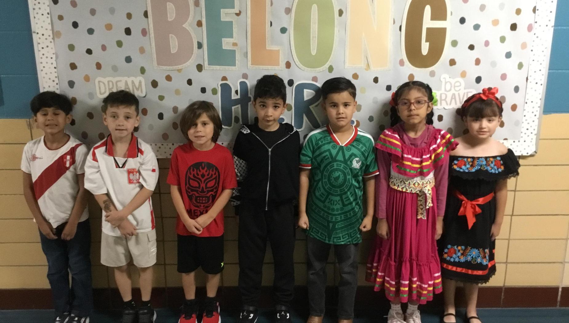 Children in diverse cultural attire stand in front of a colorful 'Belong Here' sign.
