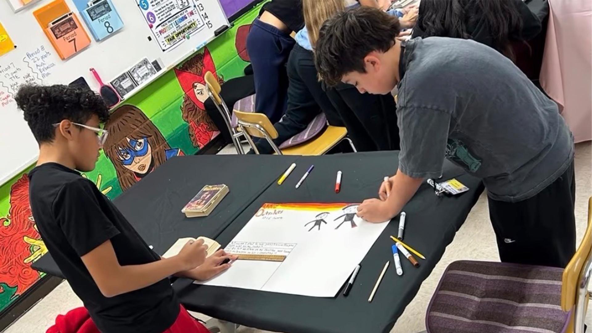 Two students drawing on a large paper at a black table, with others working in the background.
