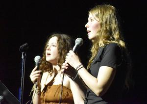 Two young women sing together with microphones on stage.