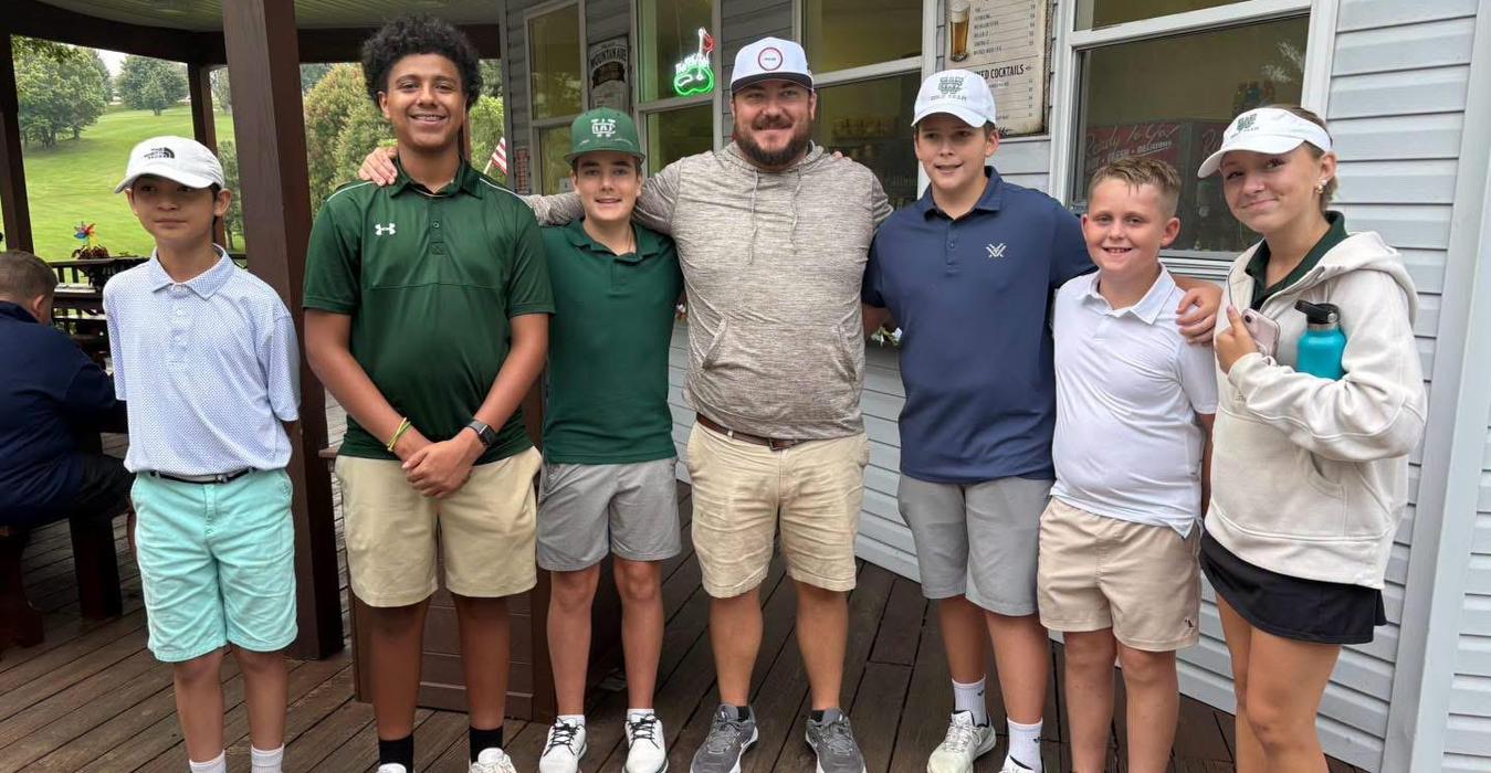 Seven young golfers and a coach pose together outside a clubhouse.