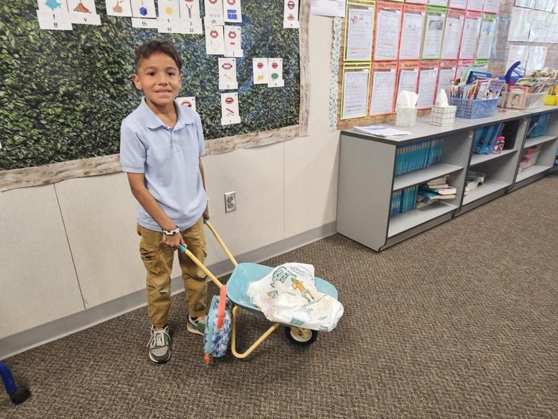 A child pushing a toy wheelbarrow inside a classroom with educational materials.