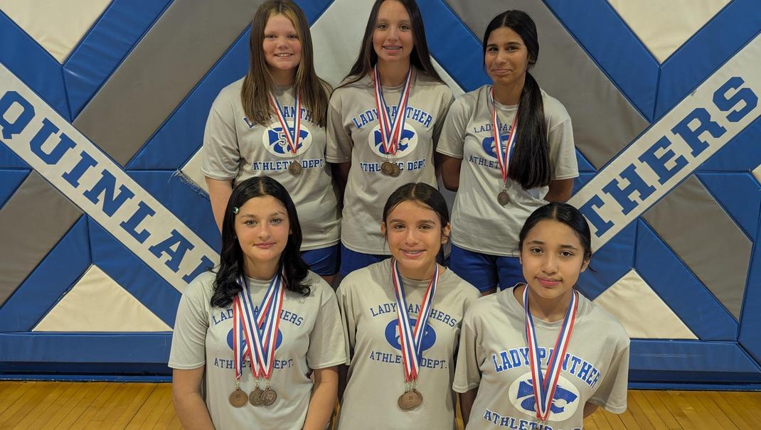 Six female athletes wearing medals and matching shirts pose in front of a gym backdrop.