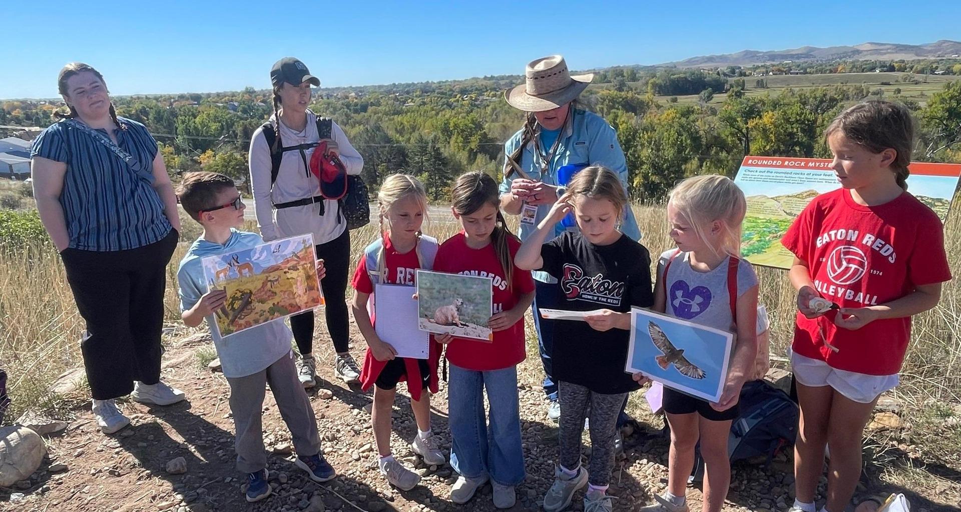 A group of children and adults on a hiking outing, engaged in nature education with photos.