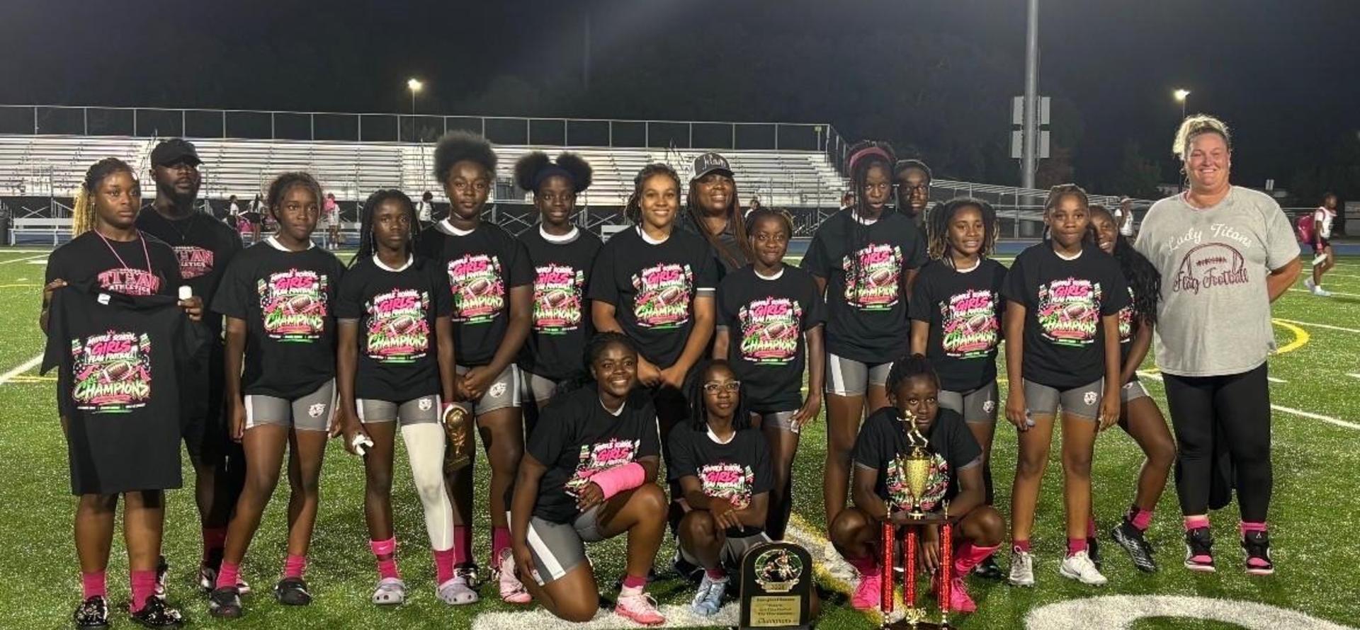 Lady Titans Undefeated Flag Football Team 2026 celebrating with trophies on a field at night.