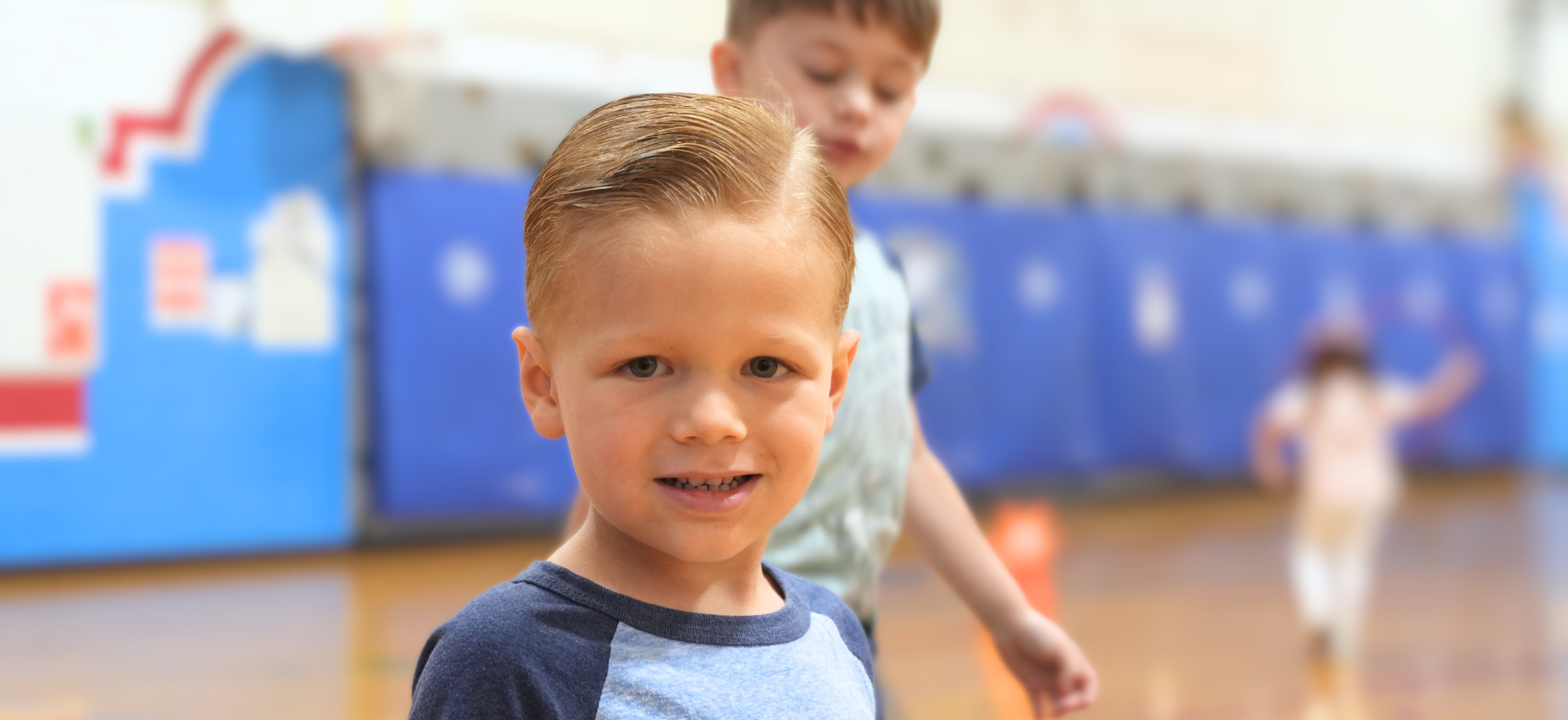 Smiling boy with styled hair in a gym, with other children in the background.