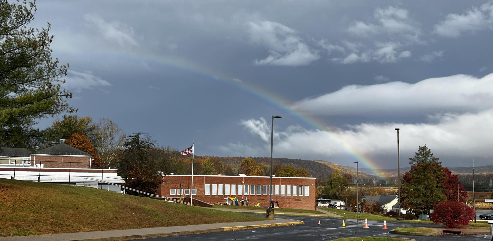A school building with a rainbow in the background and trees in autumn colors.