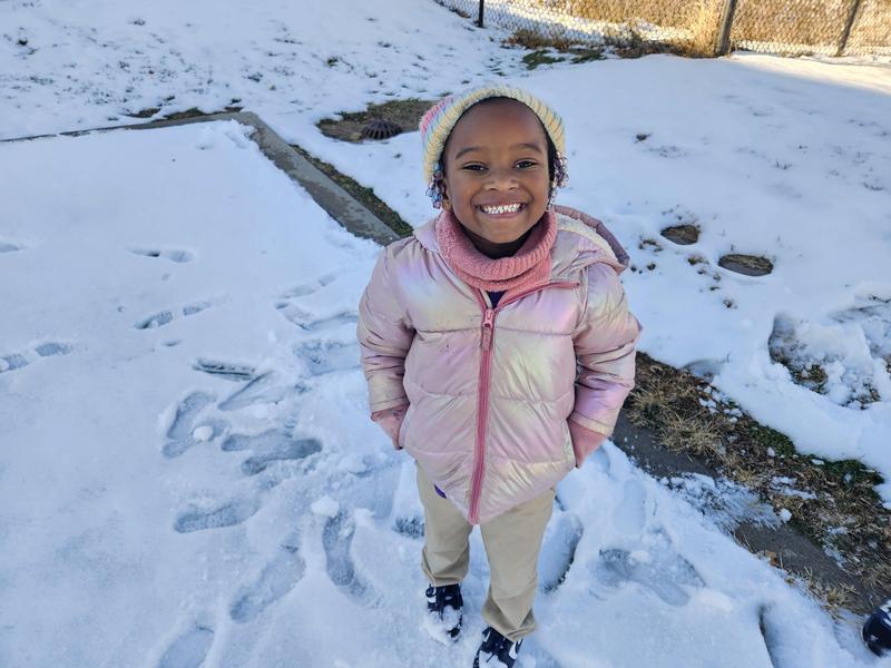A scholar stands outside in the snow in a pink coat and white hat