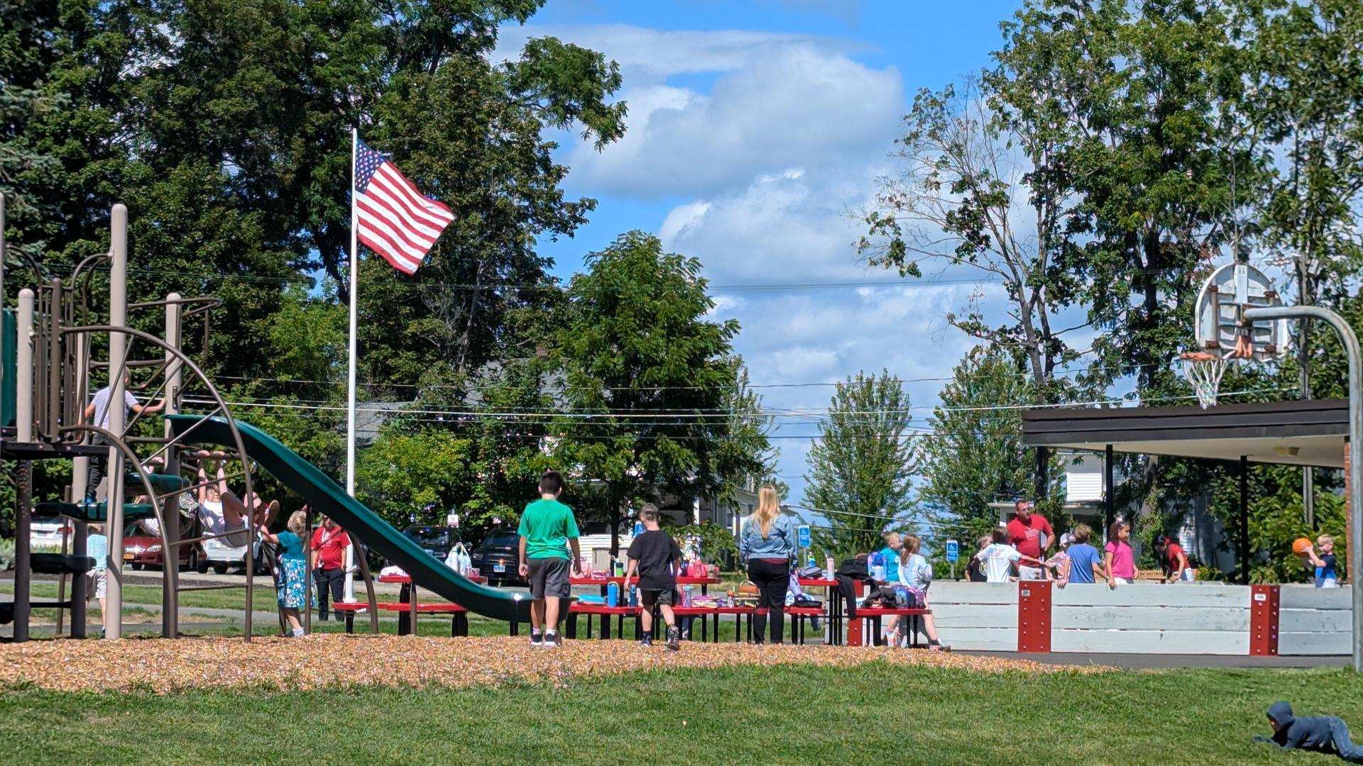 Children enjoying recess with a slide, play scape, and an American flag in the background.