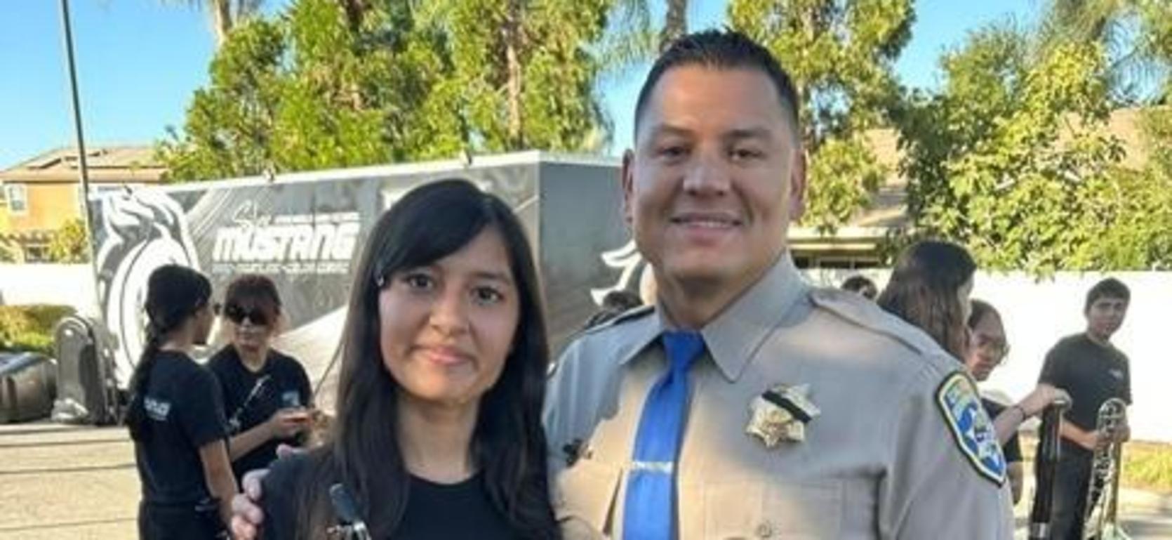 A musician holding a clarinet stands next to a police officer, both smiling together.