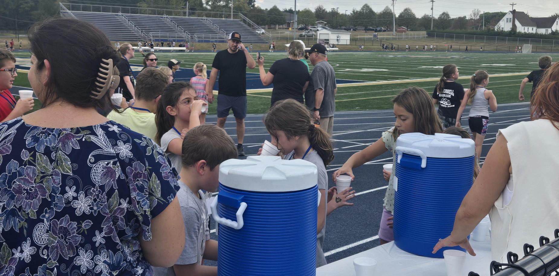 Crowd of children and adults gathering around beverage coolers at a sports event.