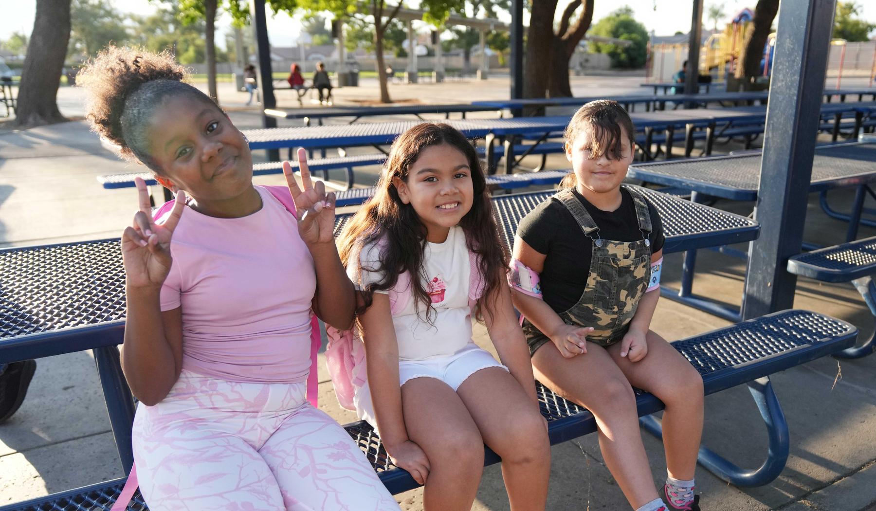 Three children sitting on benches in a school playground, smiling and posing.