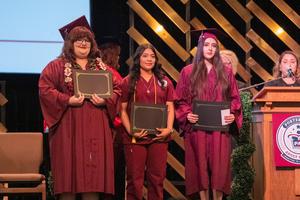 Three graduates stand proudly on stage holding certificates—two in maroon gowns and one in medical scrubs—while staff members applaud behind them.