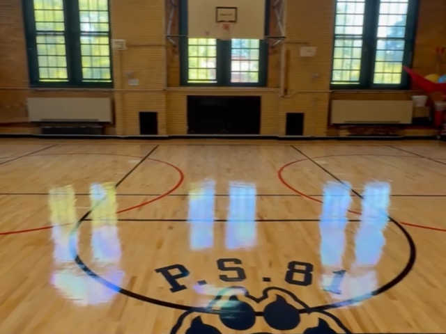 A gymnasium floor marked with a logo and basketball court lines, with sunlight streaming in.