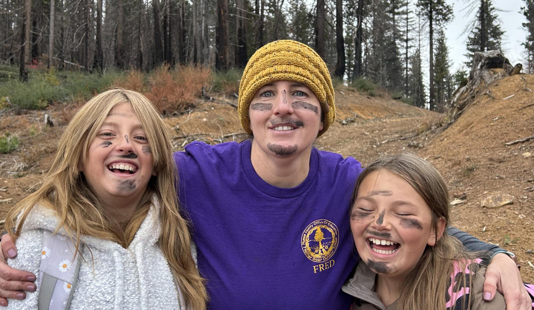 2 students flank their teacher, all are laughing outside while wearing dirt on their faces as part of an outdoor education program.