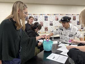 Students engaged in a classroom activity, exchanging items with a blue bucket on a table.