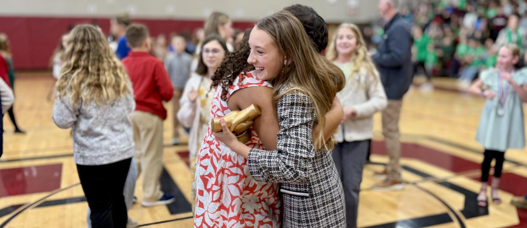 two girls smile and hug in gym with one holding trophy in her suit jacket, other in a floral dress