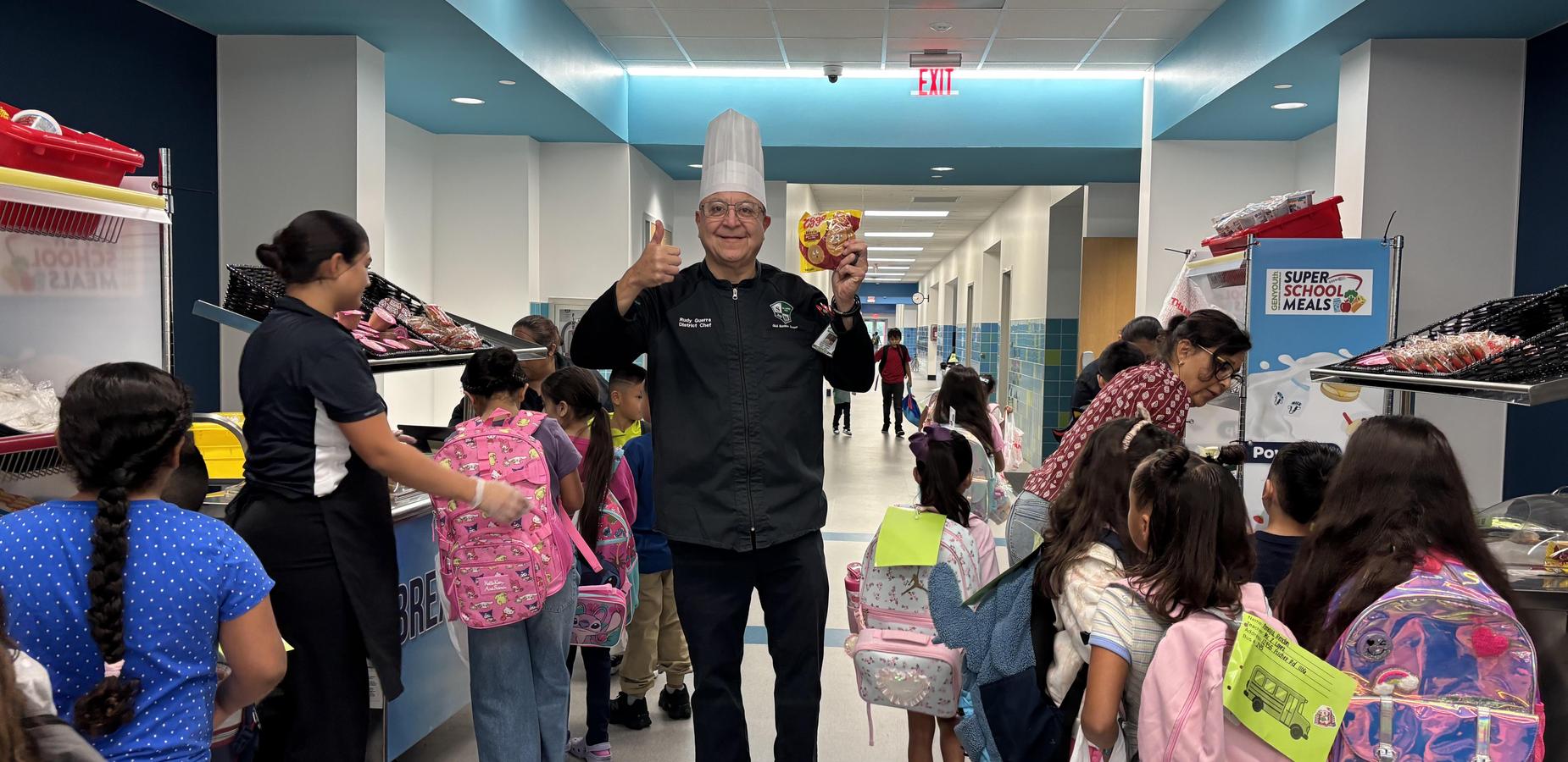 A chef holding food stands in a hallway filled with students carrying backpacks.