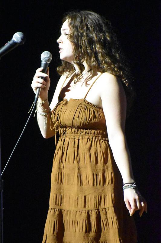 A young woman sings into a microphone, wearing a brown dress.
