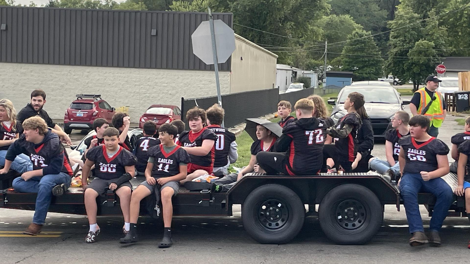 A group of young football players in jerseys sitting on a trailer during a parade.
