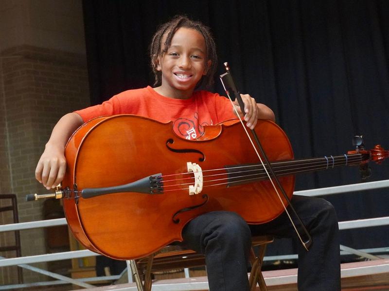 A boy seated with a cello, smiling broadly on stage.