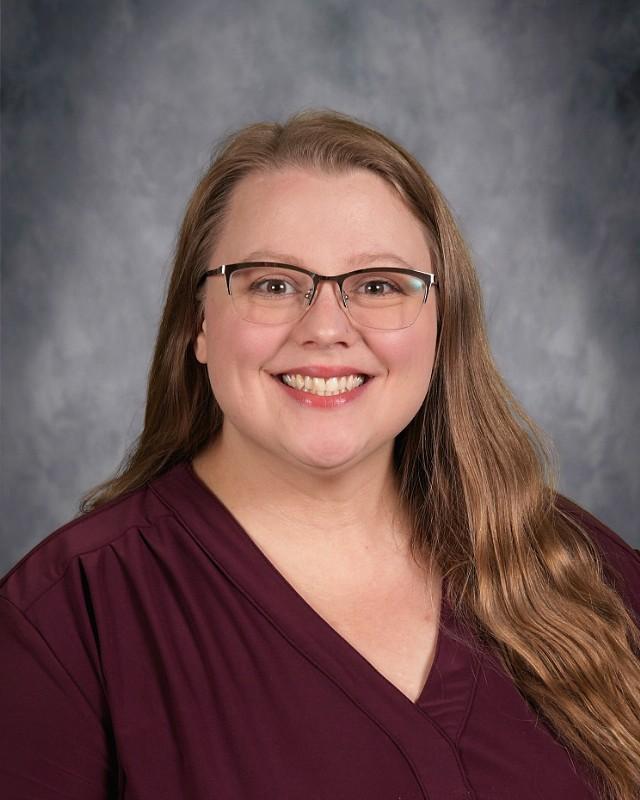 Smiling woman with long hair wearing glasses, against a grey background.