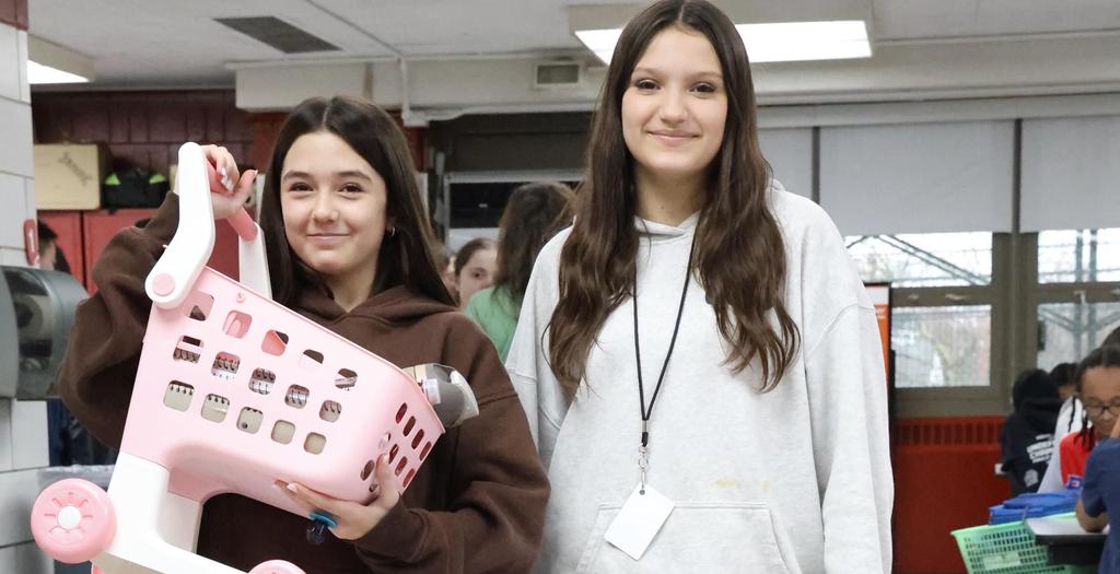 Two girls standing in the cafeteria. One girl is holding a pink toy shopping cart.