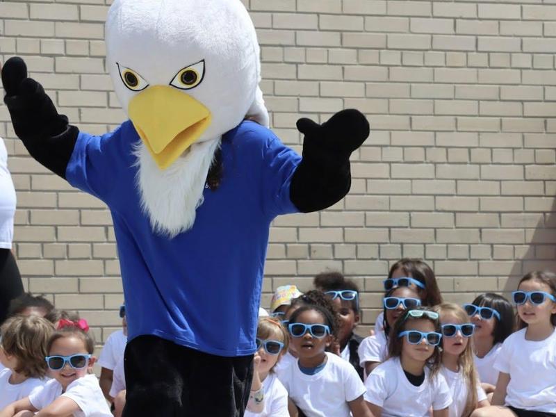 Eagle mascot dancing in front of seated children wearing sunglasses, on a stage.