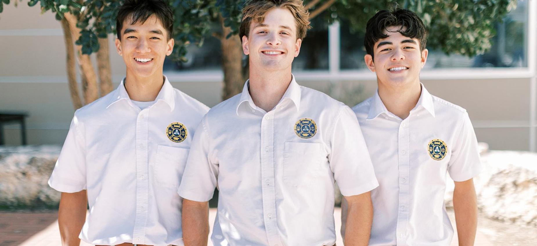 Three smiling high school students in white uniform shirts stand outside under an oak tree